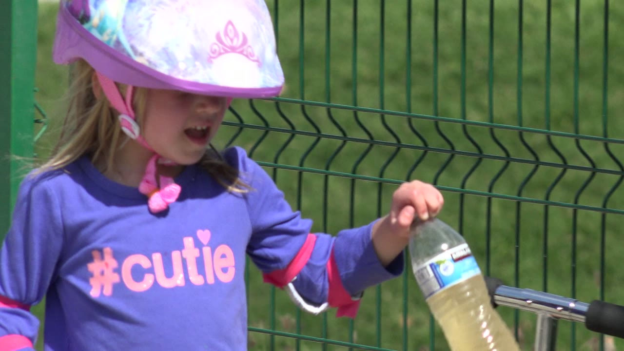 Young girl in a pink helmet holding a water bottle by a green fence.