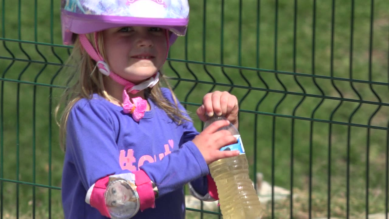 Girl in Los Cabos faces forward in a pink helmet and protective pads while gripping a water bottle against a green fence background.