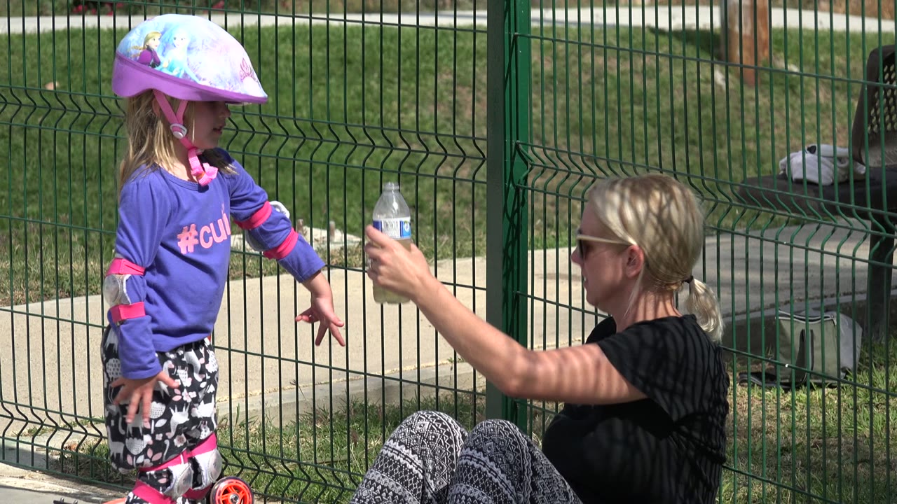 Child in a pink helmet stands near a seated woman holding up a water bottle.