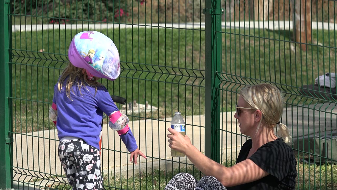 In Los Cabos, Mexico, the child faces partly away from the camera while the seated woman keeps a water bottle extended beside the park fence.