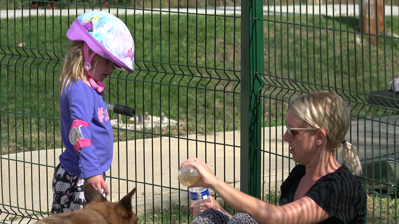 Young girl in a helmet stands beside a seated woman with water bottles near a green fence.
