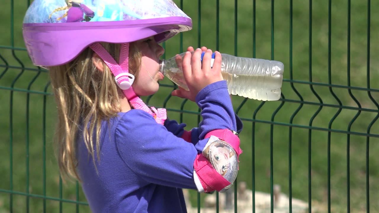 In Los Cabos, Mexico, a young girl in a purple character helmet lifts a clear plastic water bottle to her mouth beside a green metal fence.