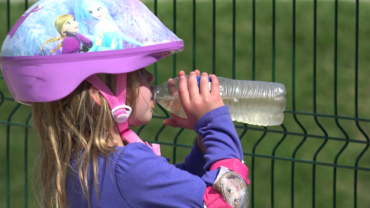 Child in a purple helmet continues drinking water near a metal fence.