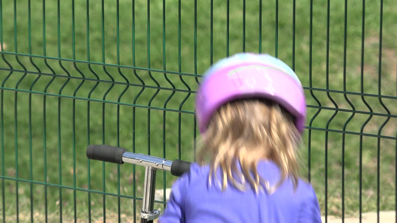 A child wearing a purple helmet stands with their back to the camera beside a scooter handlebar in a fenced park area in Los Cabos, Mexico.