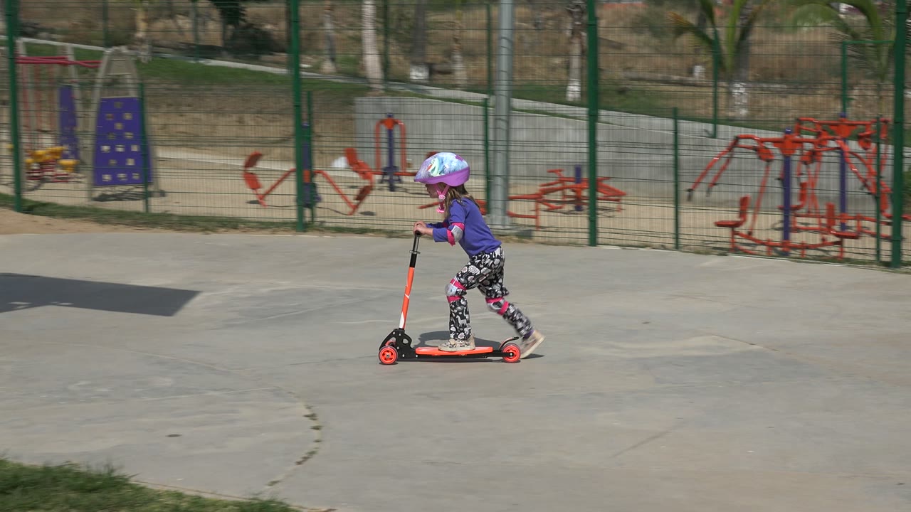 A helmeted child rides an orange scooter across a concrete path in a Los Cabos park, with playground equipment and fencing behind them.