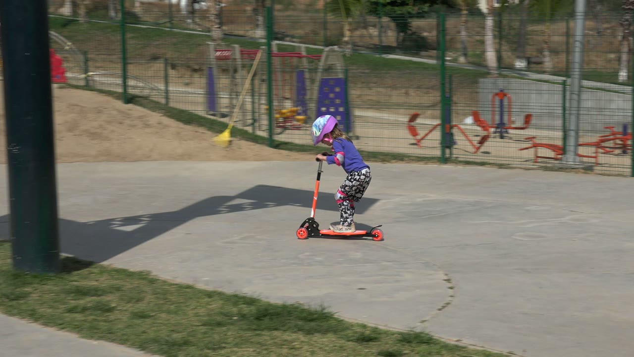 The child continues riding the orange scooter in Los Cabos, positioned near the shadow of a tall green post on the concrete path.