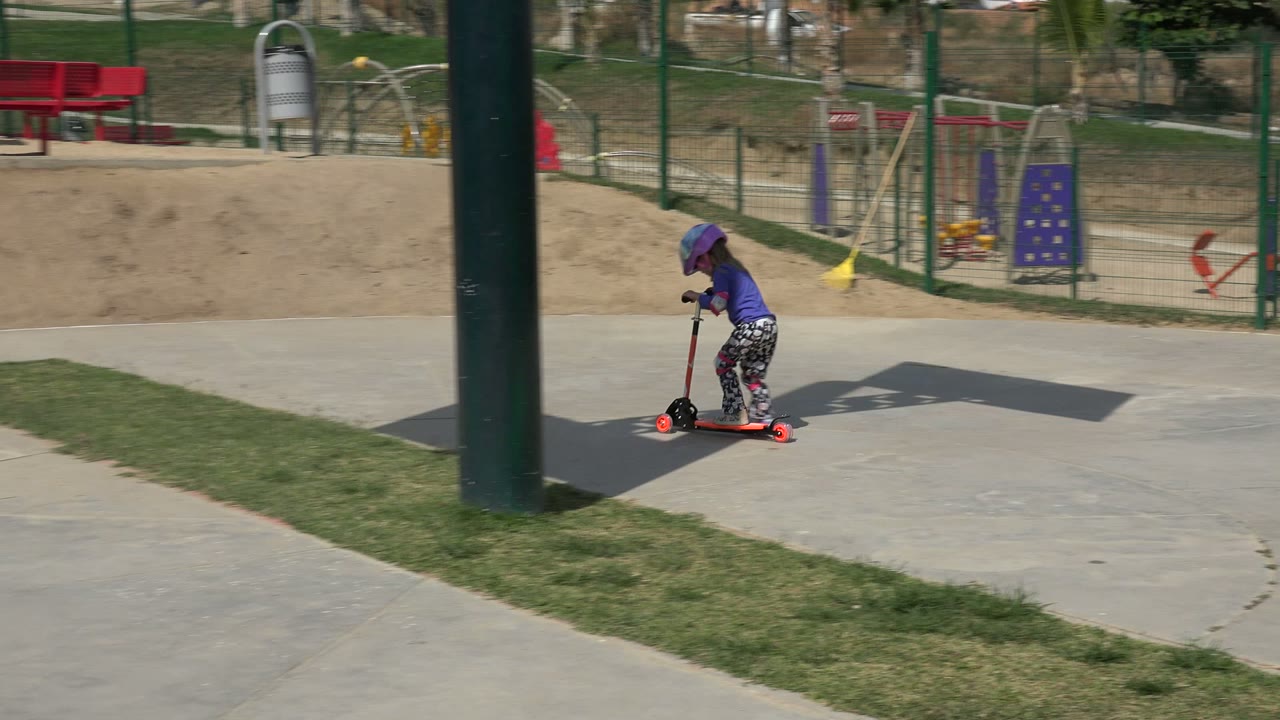 Child riding scooter near grass and playground fencing.