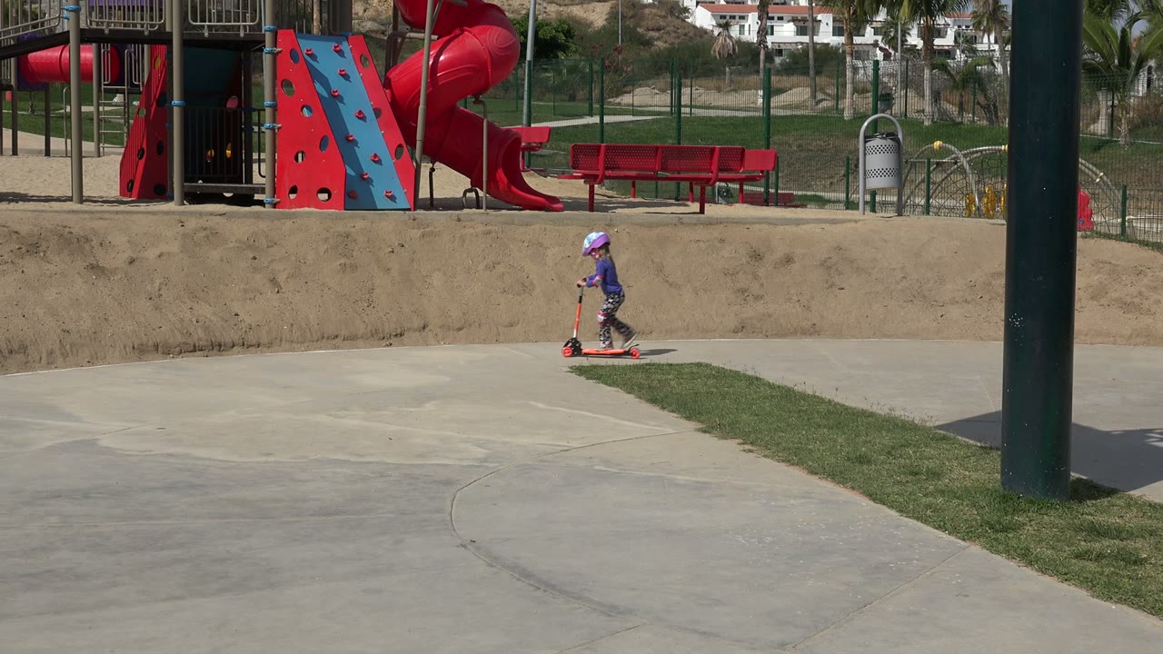 Young girl on a red scooter near playground equipment.