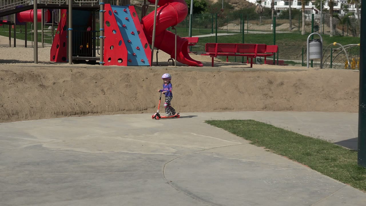 The girl continues riding the red scooter in Los Cabos, Mexico, with a dark pole in the foreground and playground slides and benches behind her.