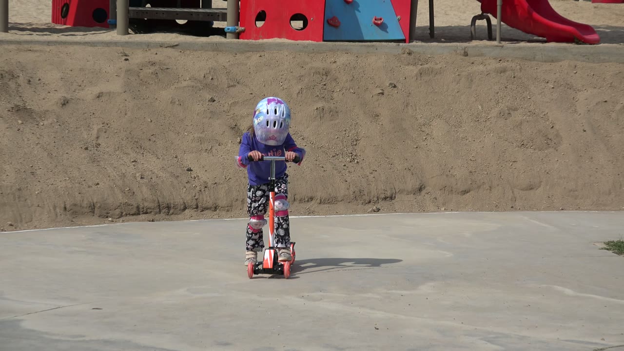 A young girl in a purple top and helmet rides an orange scooter toward the camera on a concrete park surface in Los Cabos, Mexico.