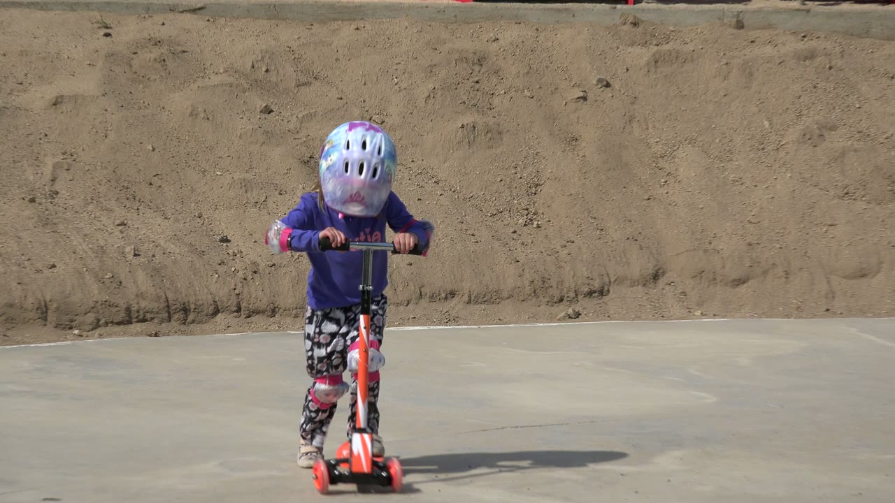 The child moves closer on the orange scooter, wearing a reflective helmet, purple top, patterned leggings, and pink pads in a Los Cabos park.