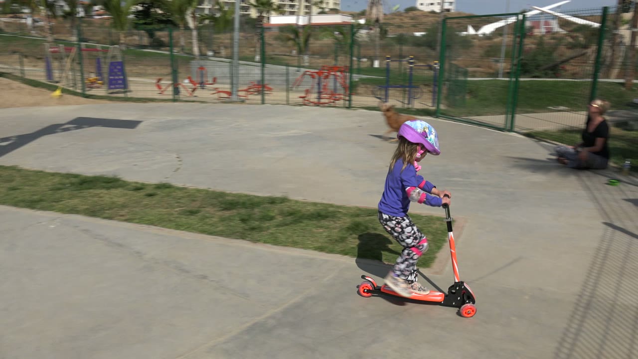 The child continues riding the scooter through the Los Cabos park, moving across the paved foreground with red playground equipment and palm trees visible beyond the fence.