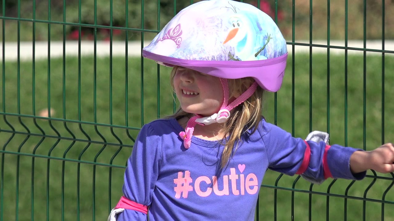 The child in Los Cabos, Mexico smiles under a purple bike helmet while holding the green fence, with soft grass and a path in the background.