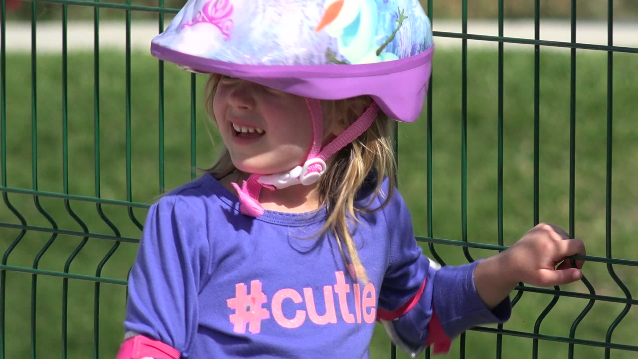 Young girl in Los Cabos wearing a pink printed bike helmet and purple shirt, standing close to a green metal fence in bright sunlight.