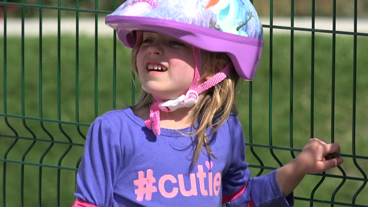 Smiling young girl in Los Cabos wearing a colorful helmet, with one hand on the green fence and sun lighting her face and shirt.
