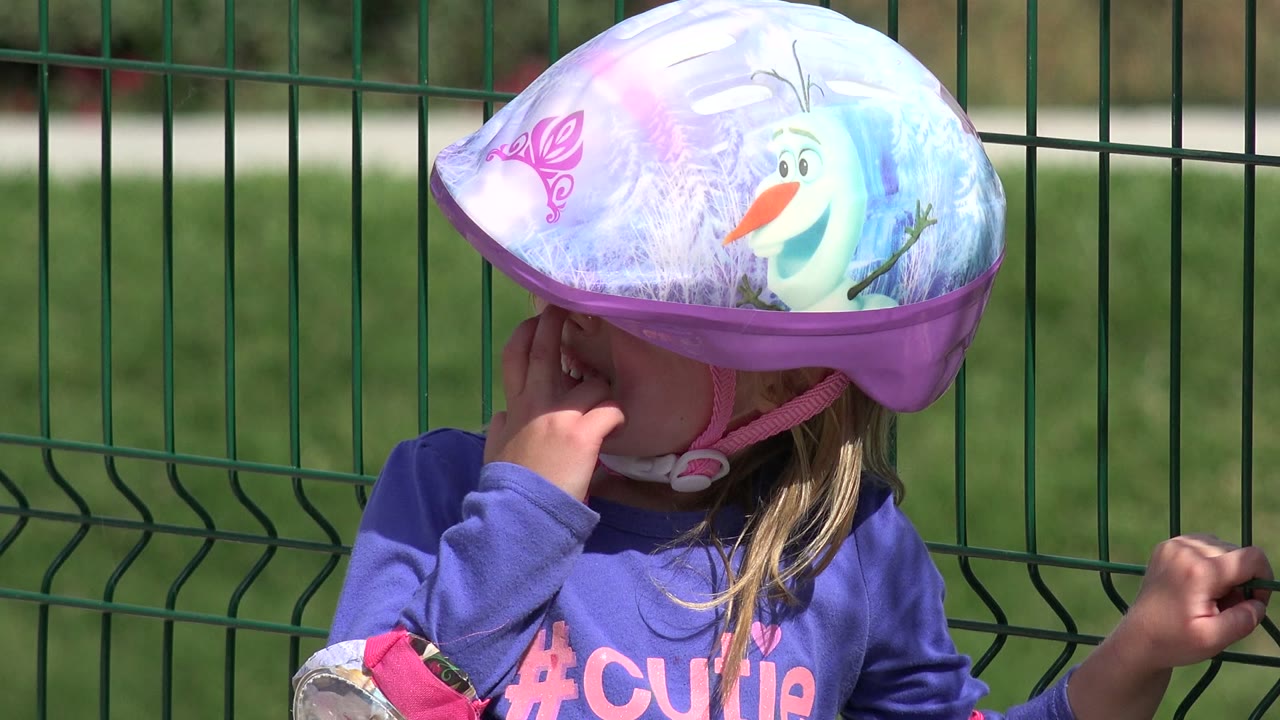 Young girl in a purple helmet stands by a green metal fence.