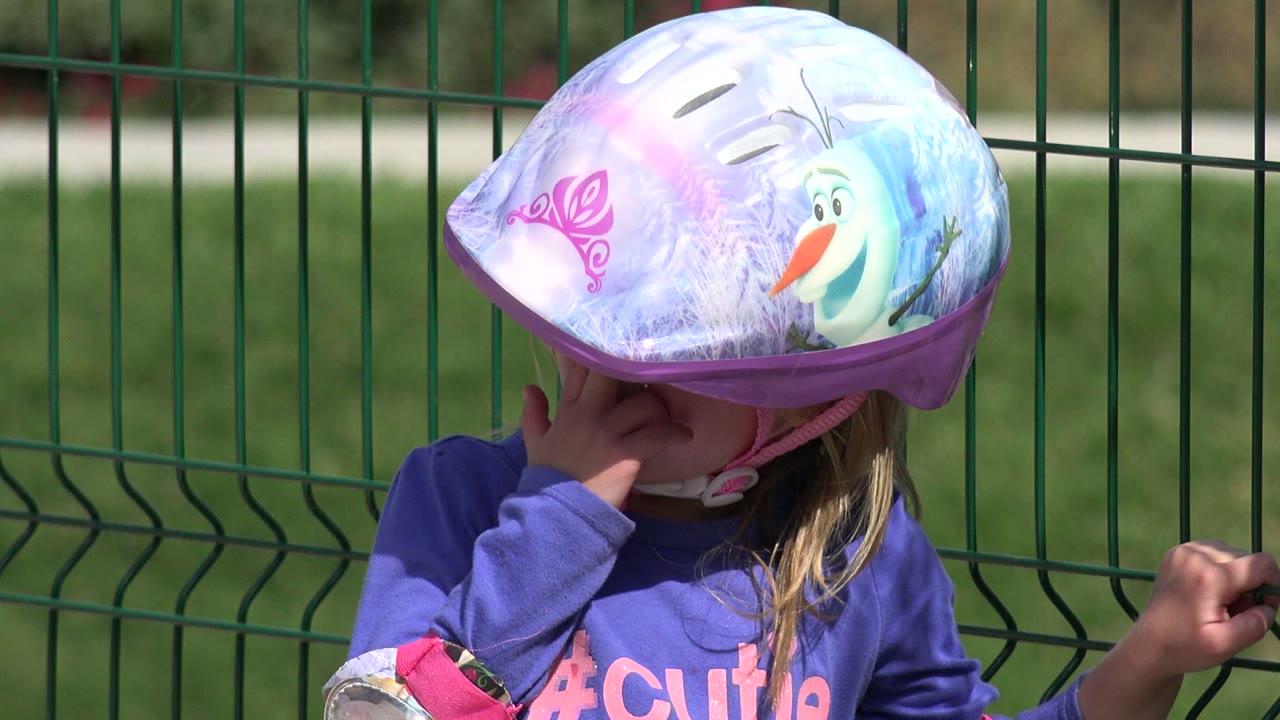 At 0:01 in Los Cabos, Mexico, the young girl remains beside the green fence with her helmet low over her eyes and her hand still touching her face.