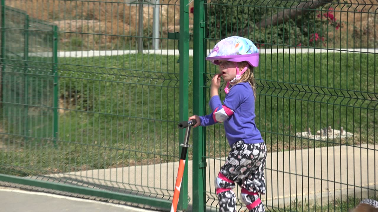 A young child in a colorful helmet and pads holds a scooter near green fencing in a Los Cabos playground, with grass and a concrete path behind.