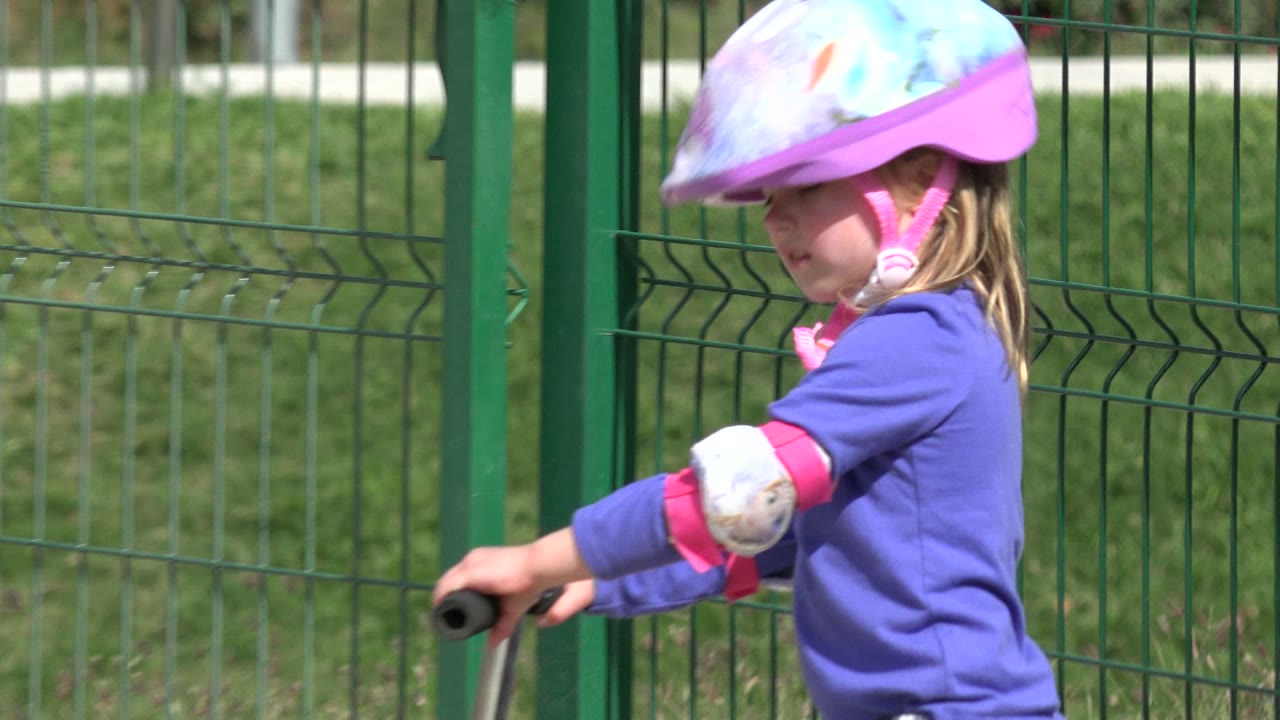 A close side view shows the helmeted child riding the scooter along green fencing in Los Cabos, with bright sun on the purple shirt and safety pads.