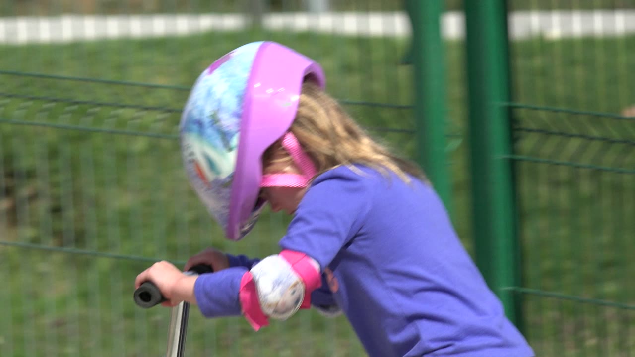 Child leaning over scooter handlebars beside a green fence.