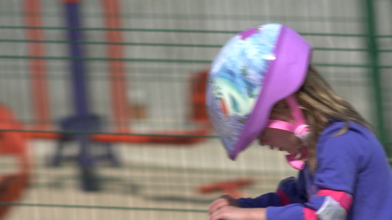 At the start of the Los Cabos clip, a young girl in a purple helmet and shirt rides in side profile with a green fence and blurred park seating behind her.