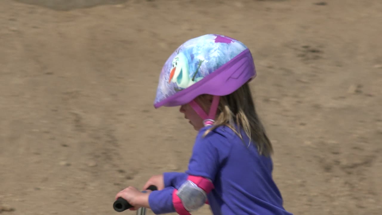Girl in a purple shirt rides a scooter past a dirt bank.