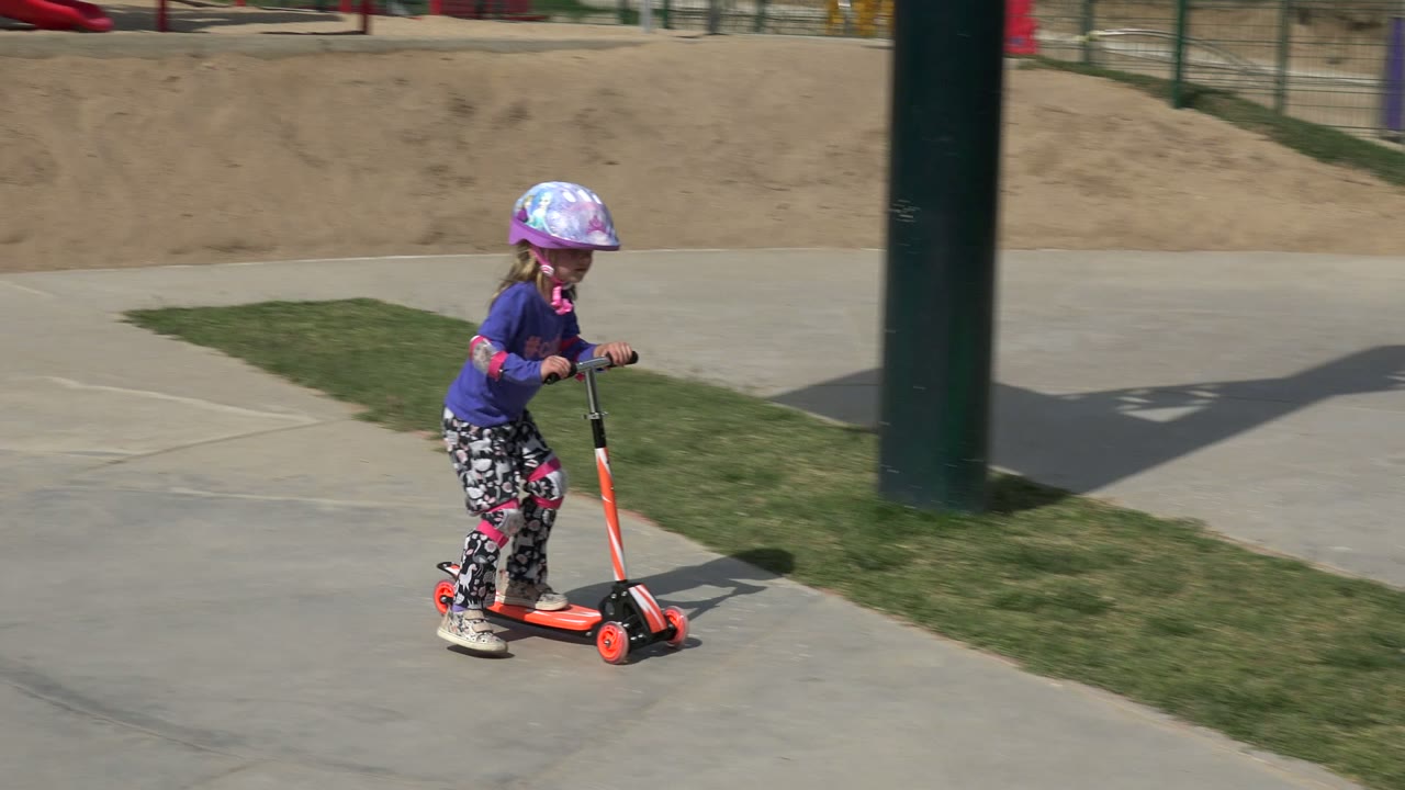 The child continues riding the orange scooter in Los Cabos, Mexico, passing a grass strip and a dark vertical shade pole in bright daylight.