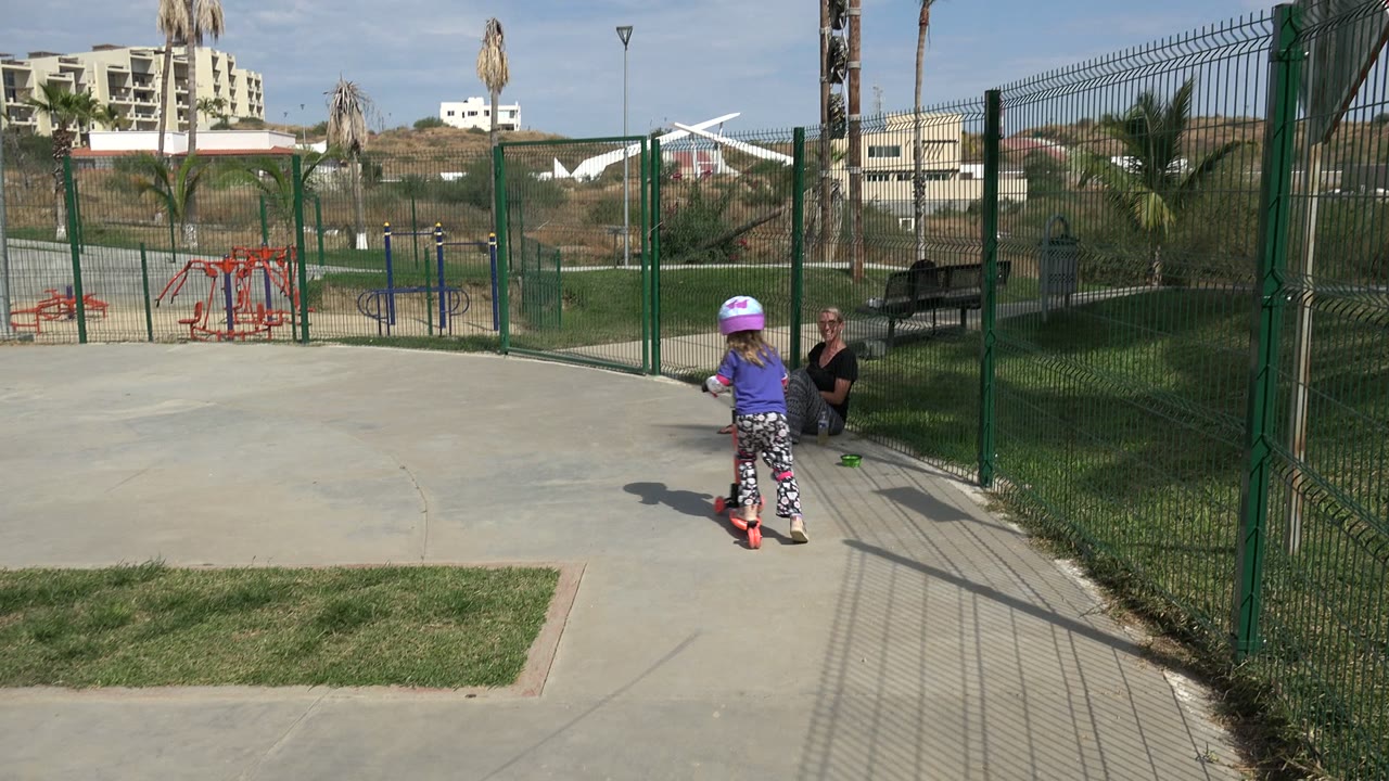Young girl on a red scooter in a fenced park with an adult seated nearby.