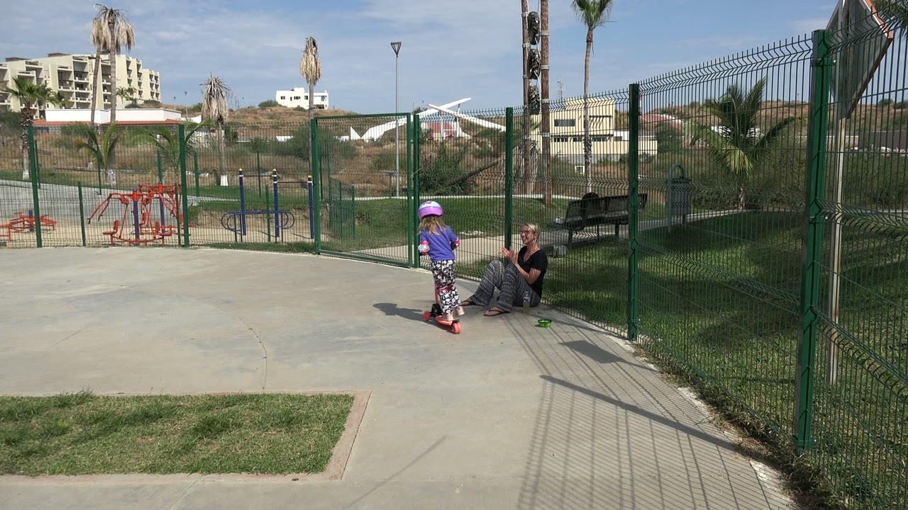 At 0:01 in Los Cabos, the helmeted girl moves farther along the concrete on her red scooter as the seated adult watches from the grass by the fence.
