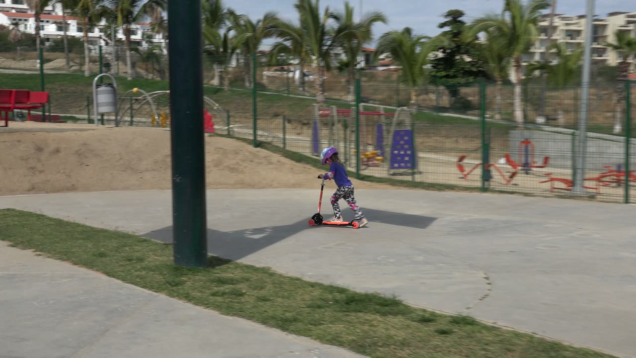 A young child in a purple helmet rides an orange scooter across a concrete path in a Los Cabos park, with grass, red benches, fencing, palms, and playground equipment behind.