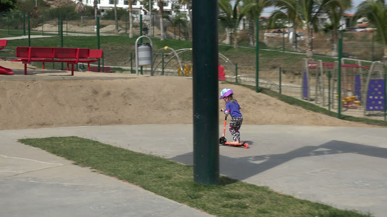 The child continues riding the orange scooter along the concrete path in Los Cabos, passing red park benches, a red slide, fencing, sand, grass, and palms in bright sun.
