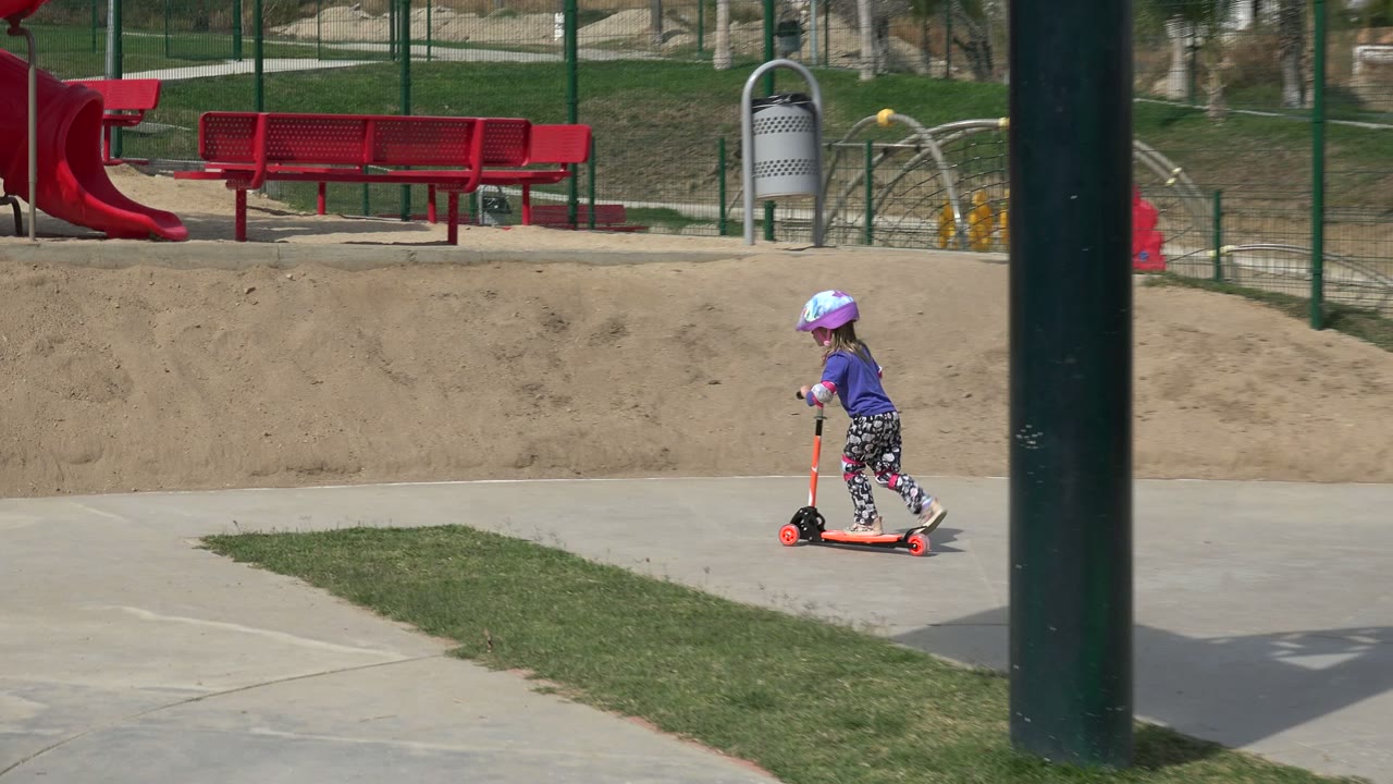 Child rides a scooter along a park path beside grass and sand.