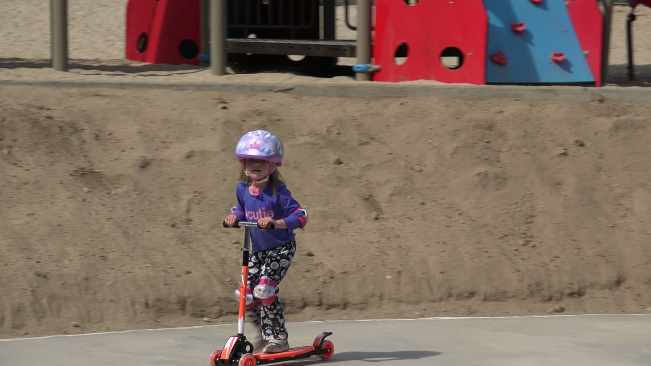 Young girl in a helmet riding an orange scooter on a park path.