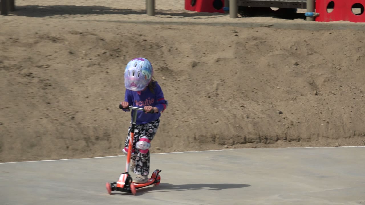 The young girl looks down while riding the orange scooter on the concrete path, with the sandy slope and playground structures still visible behind her in Los Cabos, Mexico.
