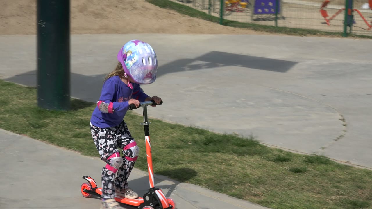 Young girl in a purple helmet rides an orange scooter beside grass.