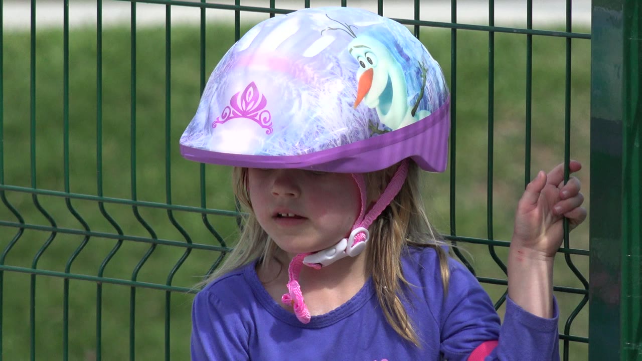 Young girl in Los Cabos faces forward under a purple Frozen-themed bicycle helmet, with the green fence filling the background.