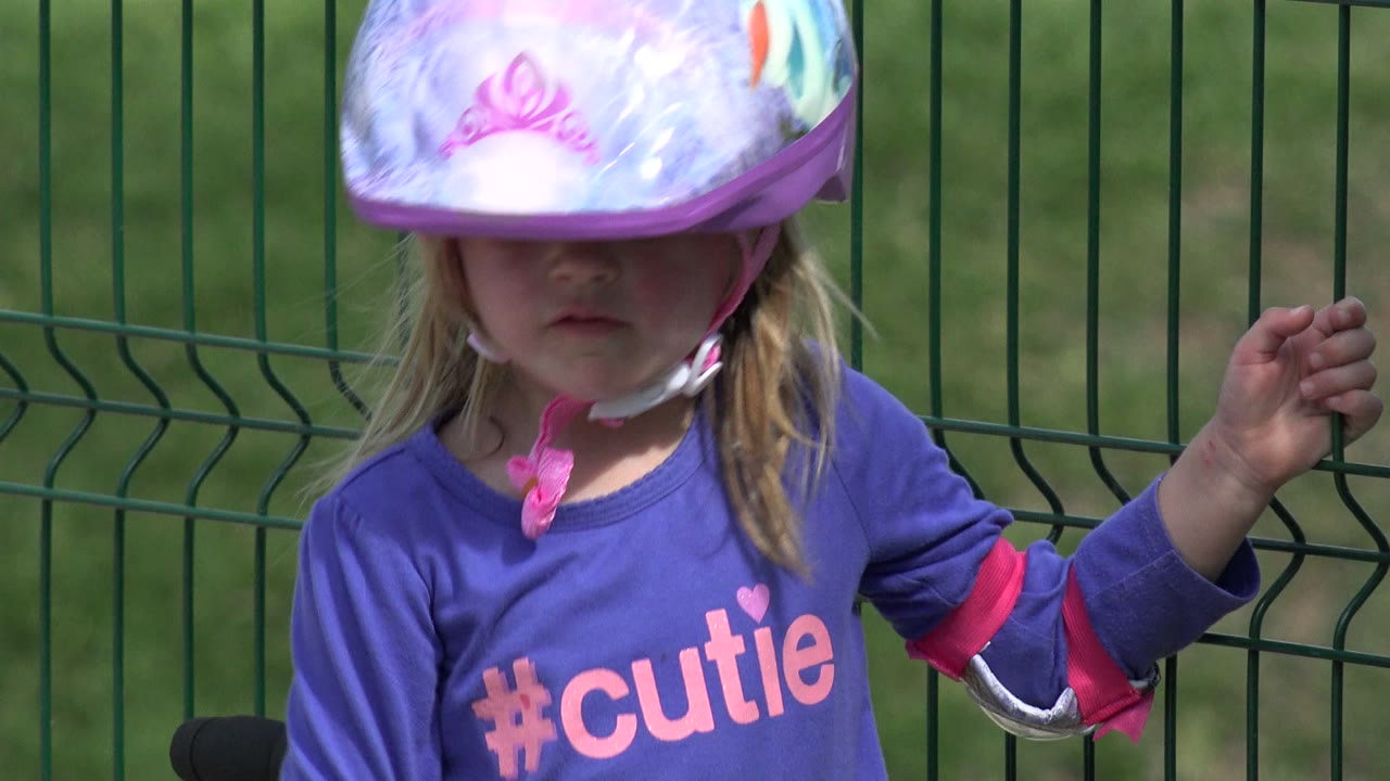 Young girl in Los Cabos, Mexico wearing an oversized purple helmet, partly shading her face as she stands next to a green metal fence.