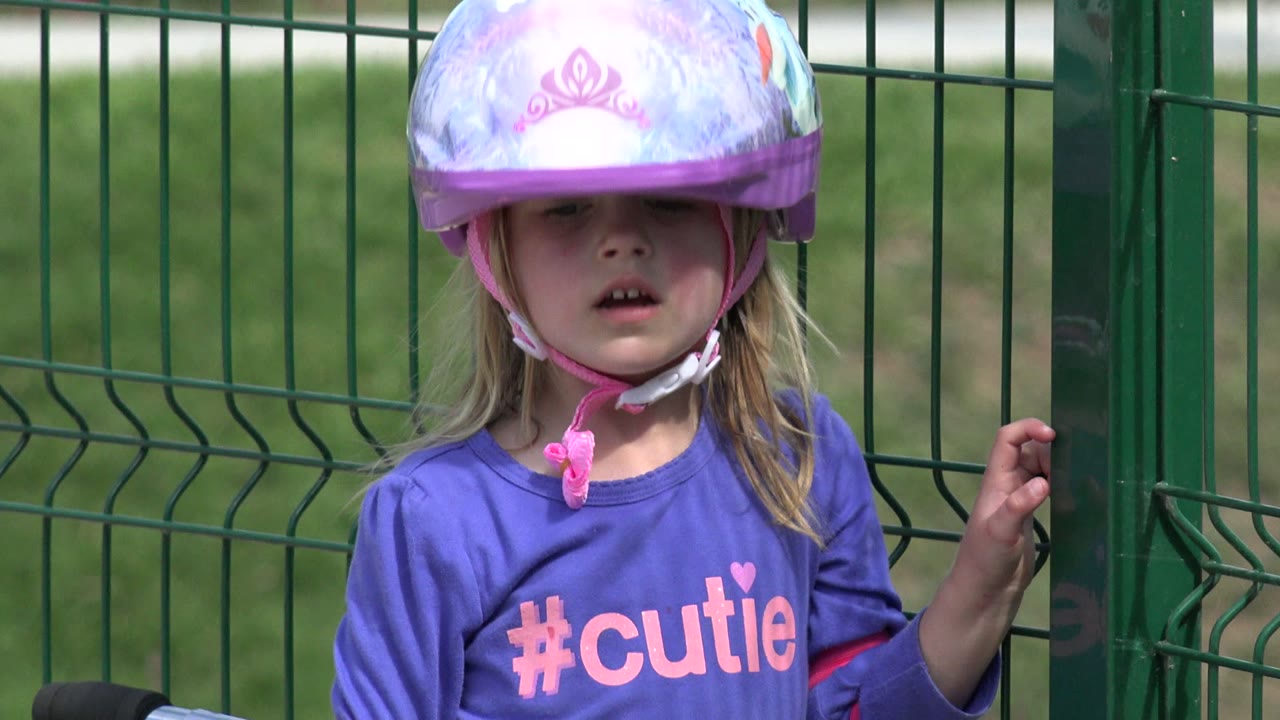Young girl in a purple shirt and helmet beside a green fence.