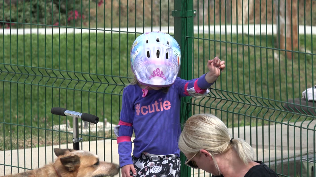 Child in helmet standing by a fence as a woman sits nearby and a dog appears at left.