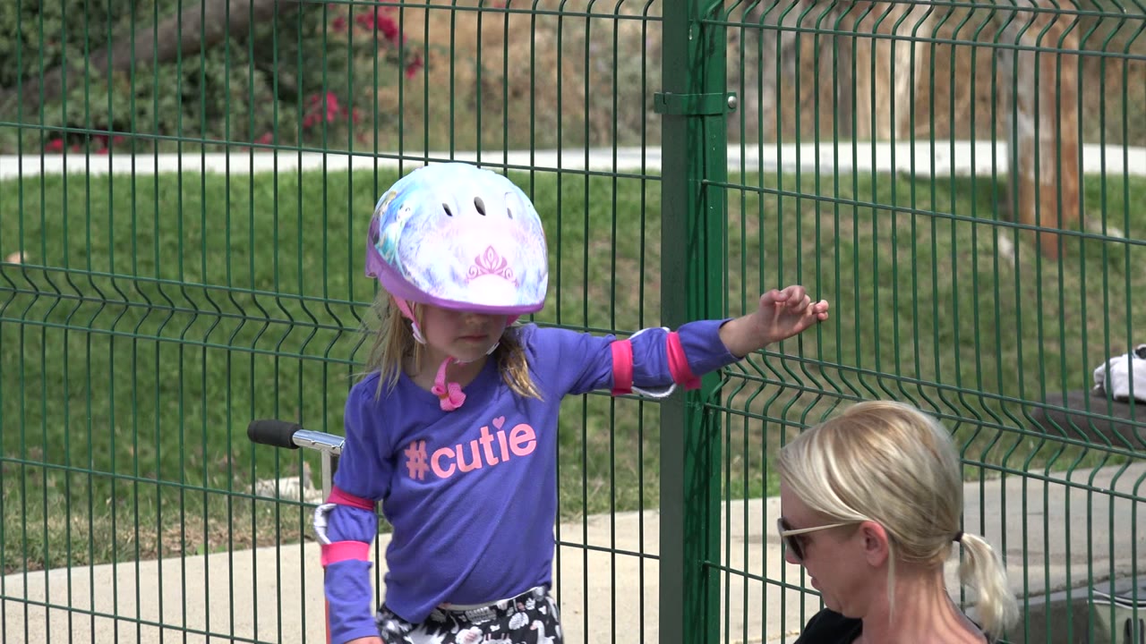 Helmeted child in purple shirt near a seated woman by a green fence.