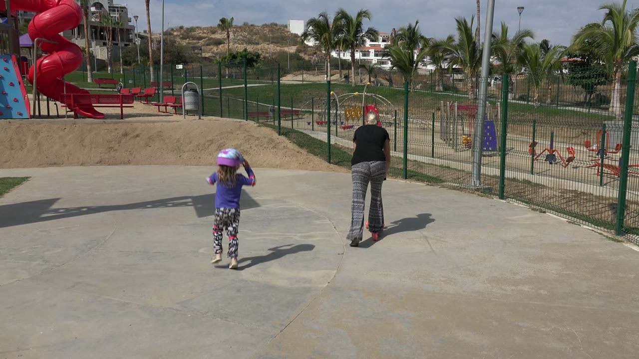 Child in a helmet and adult near a playground on a sunny concrete area.