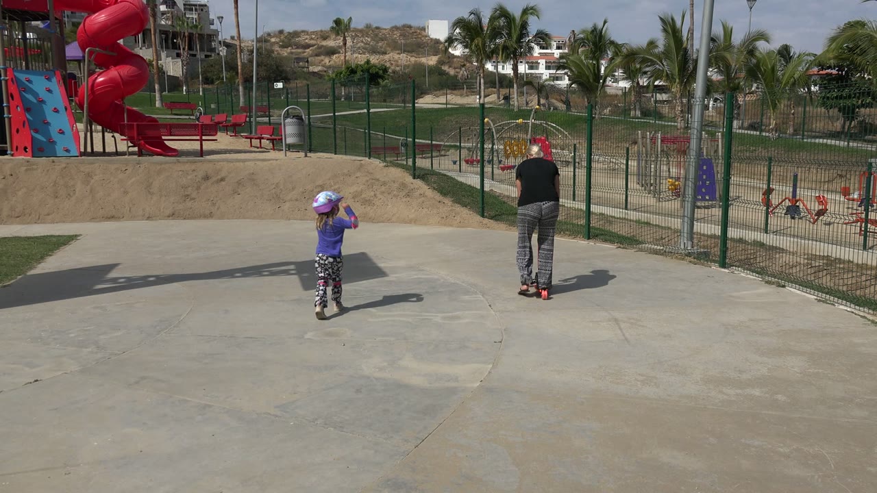 In Los Cabos, Mexico, the child stands near the center of the concrete while the adult moves away near a fence, with red slides and palms behind them.
