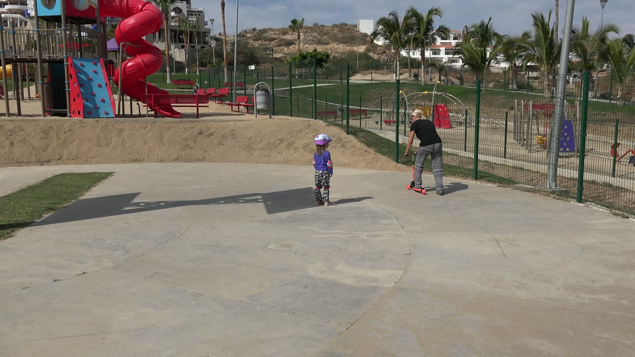 Woman with a small red scooter and a child in a purple helmet at a sunny playground park.