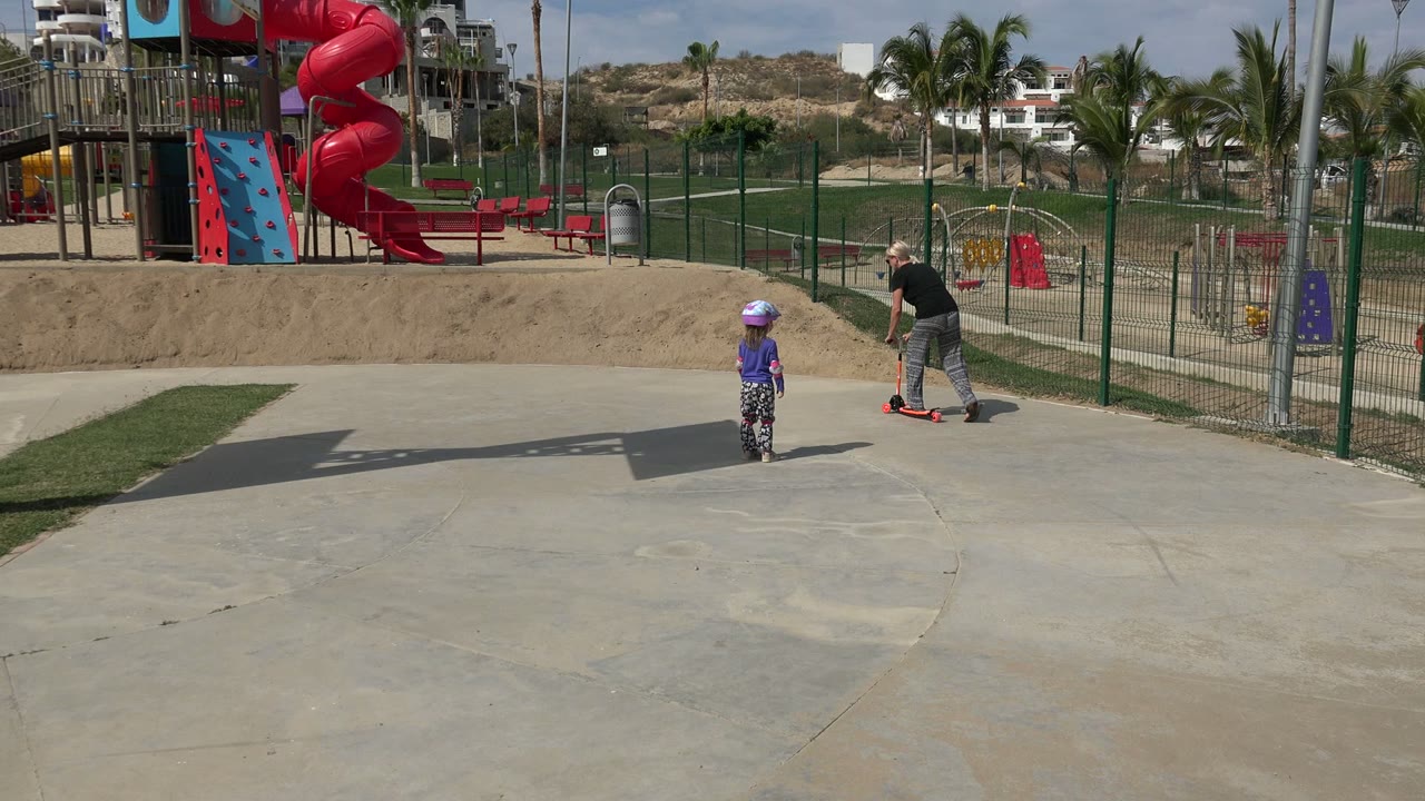 At 0:01 in Los Cabos, the child remains on the concrete while the woman bends forward over the small red scooter near the fenced playground and red spiral slide.