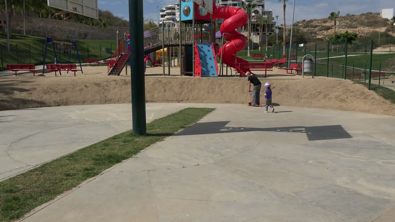 At 0:01 in Los Cabos, the father and child remain near the scooter on the concrete path, framed by a playground tower, red slide, benches, fencing, and hillside.