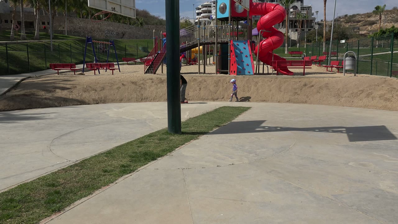 Wide Los Cabos playground frame with a small child on the paved path, red slide and swings behind, benches and palms in bright daylight.