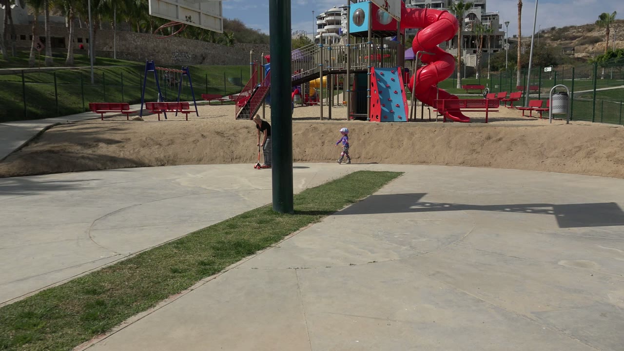 A small child moves across the Los Cabos playground pavement while the red spiral slide, blue climbing wall, benches, fence, and grass sit behind.