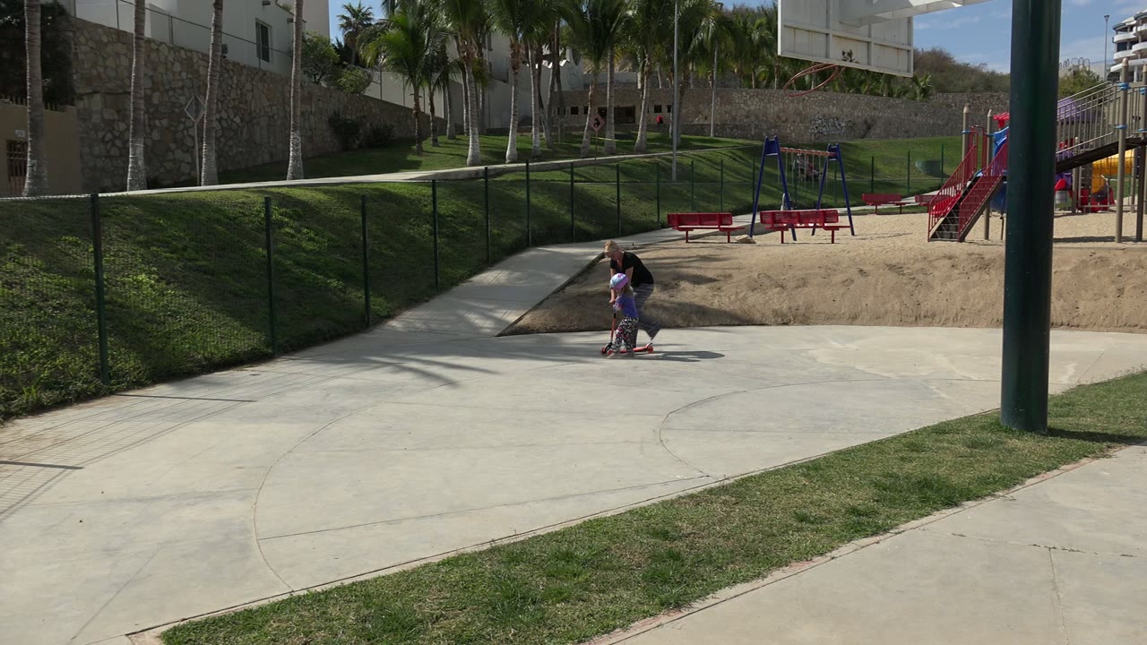 One second into the Los Cabos footage, the helmeted girl moves on the red scooter with the adult woman close beside her, framed against fencing, grass, sand, benches, and playground equipment.