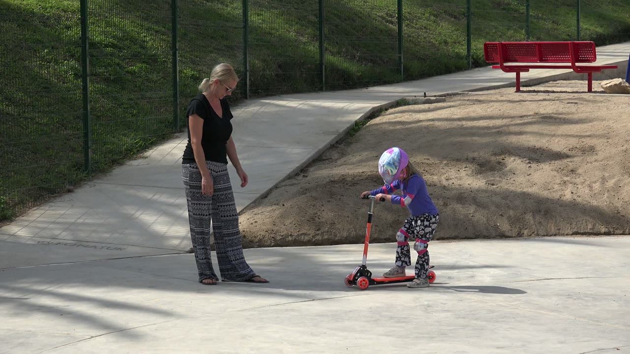 Mother stands near a child on a red scooter outdoors.