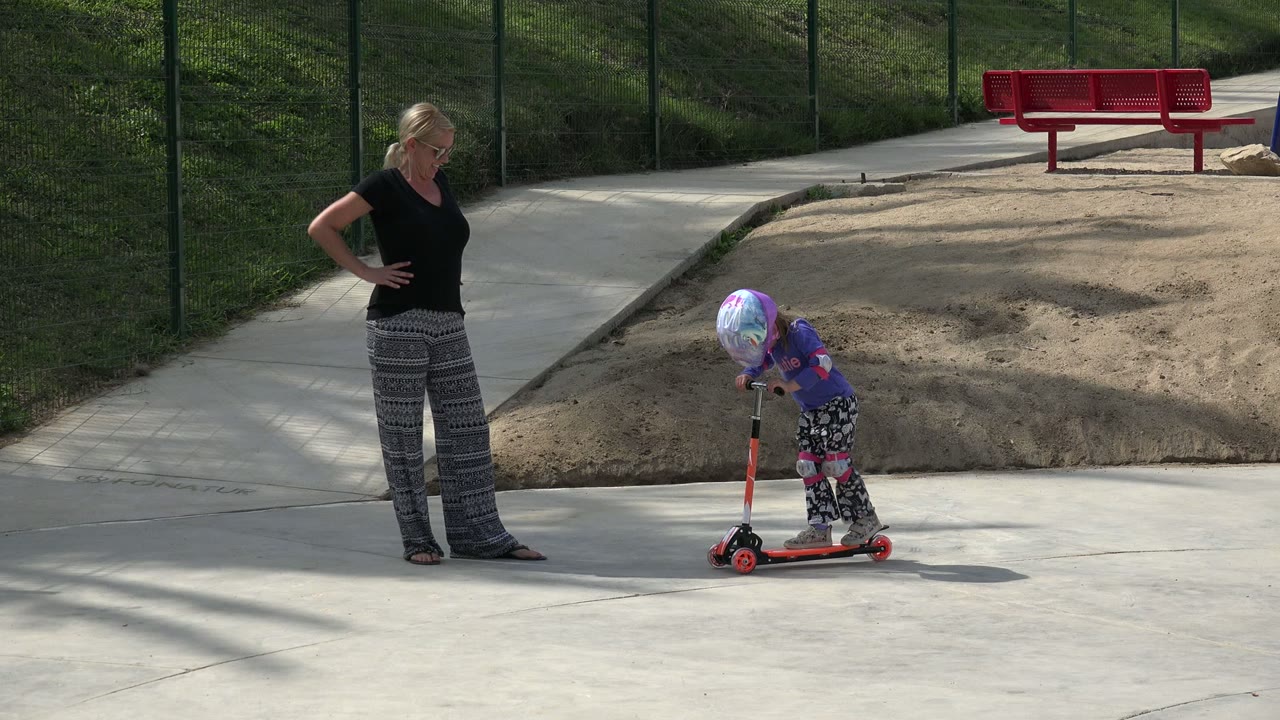 In Los Cabos, Mexico, the child bends forward over the scooter handlebars as the mother stands back with hands on hips beside the paved play area.